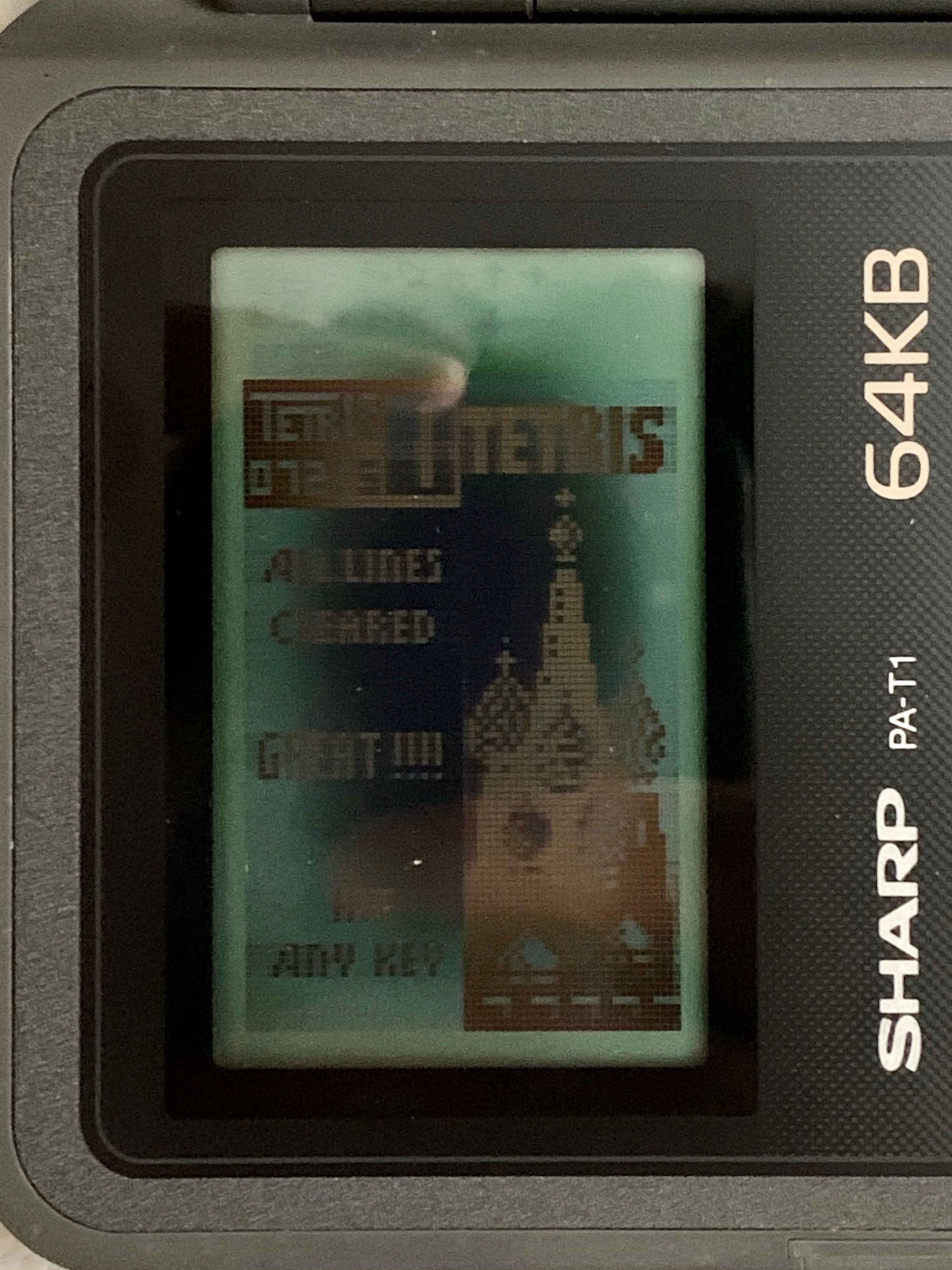 A cropped closeup of the screen of the Sharp PA-S1, the screen shows the endscreen after the Tetris game is completed. It says 'All lines cleared / Great! / Hit any key' with a pixelixed rendtion of Saint Basil's Cathedral next to the text.