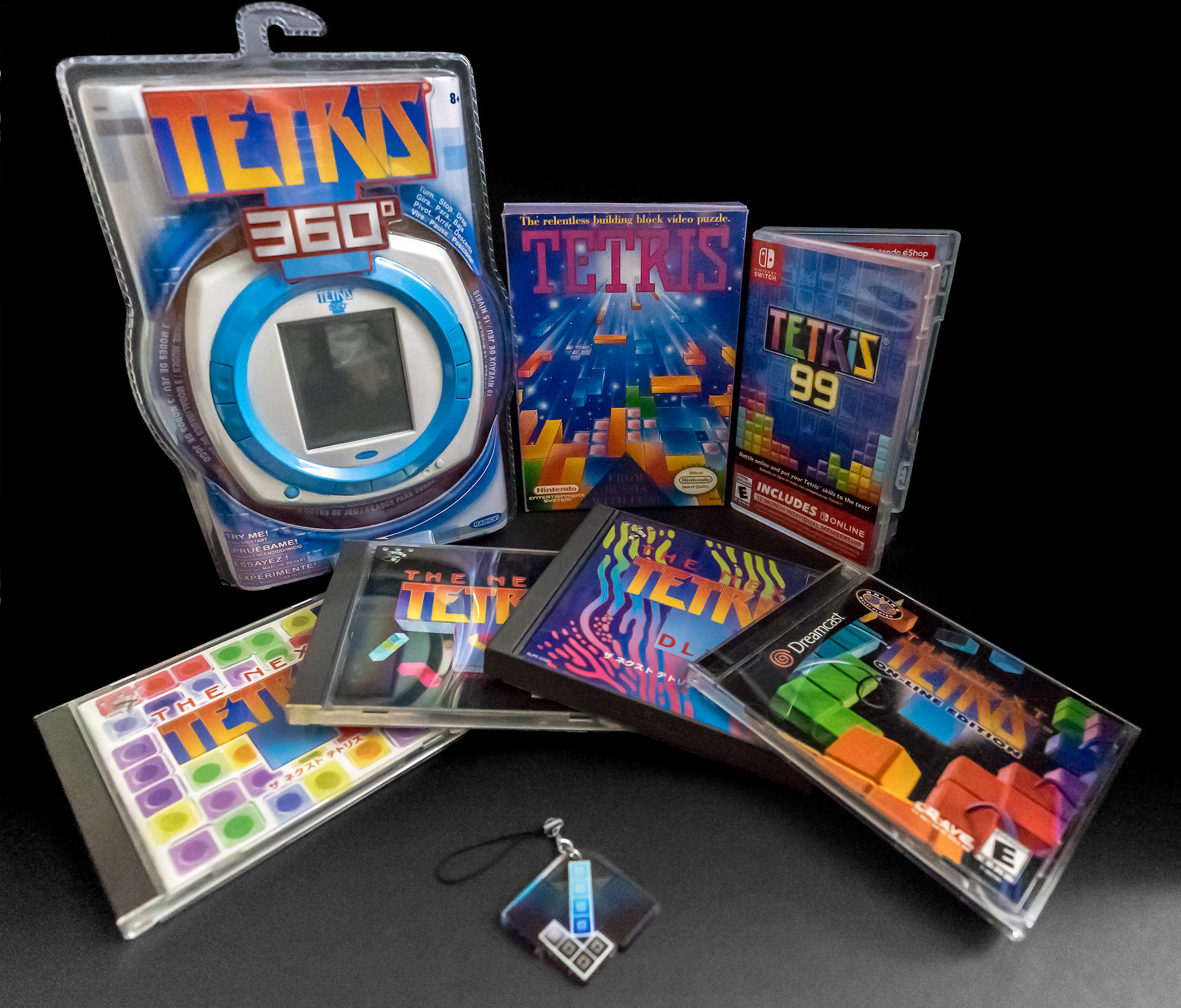 A photo showing seven Tetris related items, neatly arranged against a black backdrop. From top left to bottom right, Tetris 360, Tetris for NES, Tetris 99, three Japanese versions of the game The Next Tetris for PlayStation, The Next Tetris for Dreamcast, and a keychain made by the Hard Drop community.