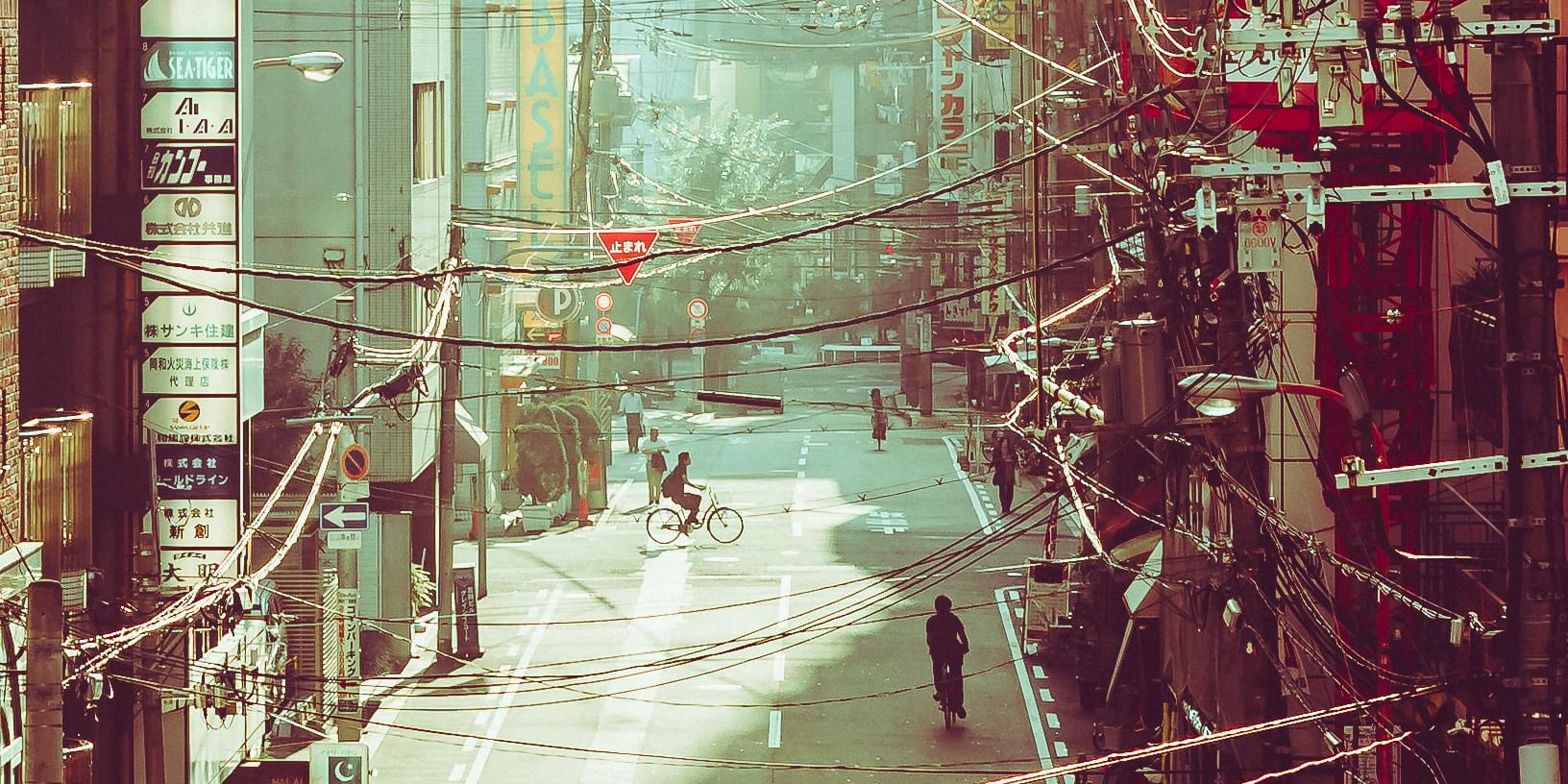 A photo taken down a street from second story vantage point, in Osaka, Japan. Buildings line the street, and power lines cross from one side to the other. Only cyclists and pedestrians can be seen on the street.