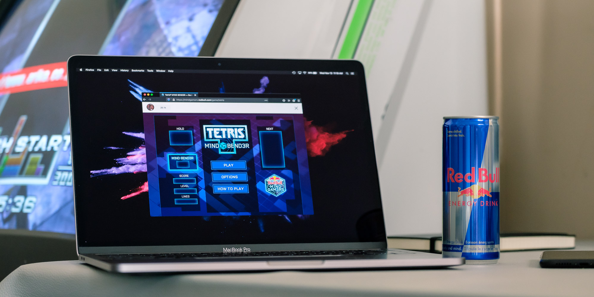 A MacBook Pro latop with the game Tetris: Mind Bender opened in windowed mode. A can of Red Bull is next to the laptop. Behind the laptop a Sega Astro City with the game Tetris: The Absolute - The Grand Master 2 PLUS is playing the attract mode.
