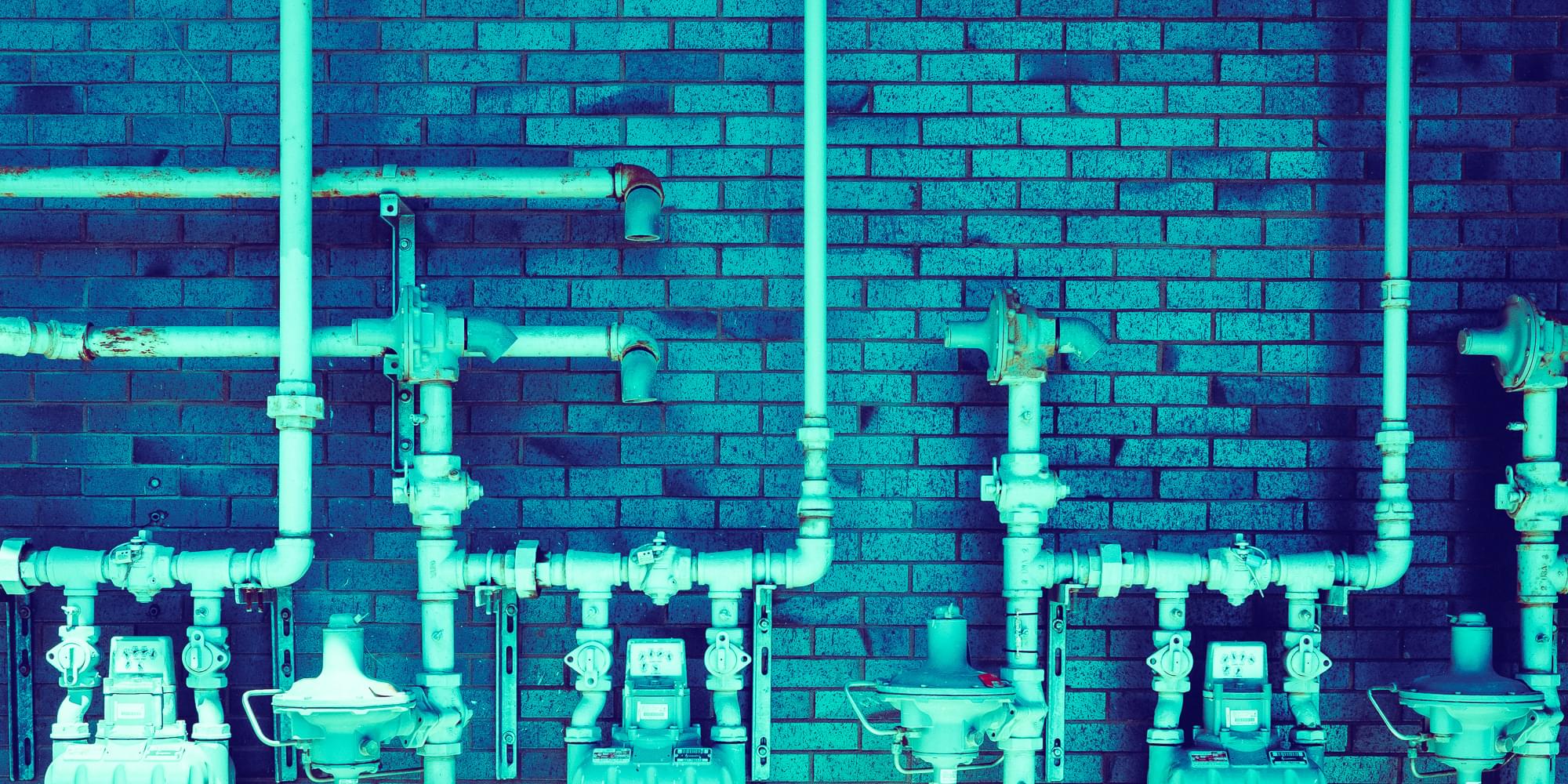 A photo of many interconnecting pipes against a backdrop of a brick wall. The photo is heavily tinted blue and cyan.
